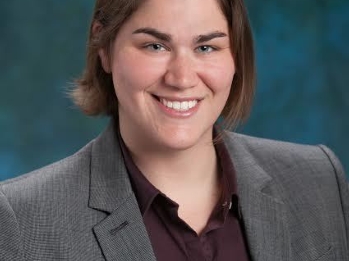 A white woman, with dark brown hair, in a gray suit jacket, dark red button-down blouse, in front of a blue-green background.
