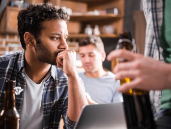 Man sitting pensively with his chin in his hand at a bar while others around him drink.