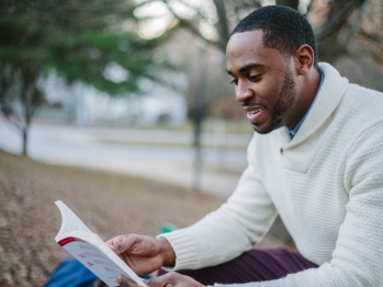 Black man wearing a white sweater sitting outside reading a book