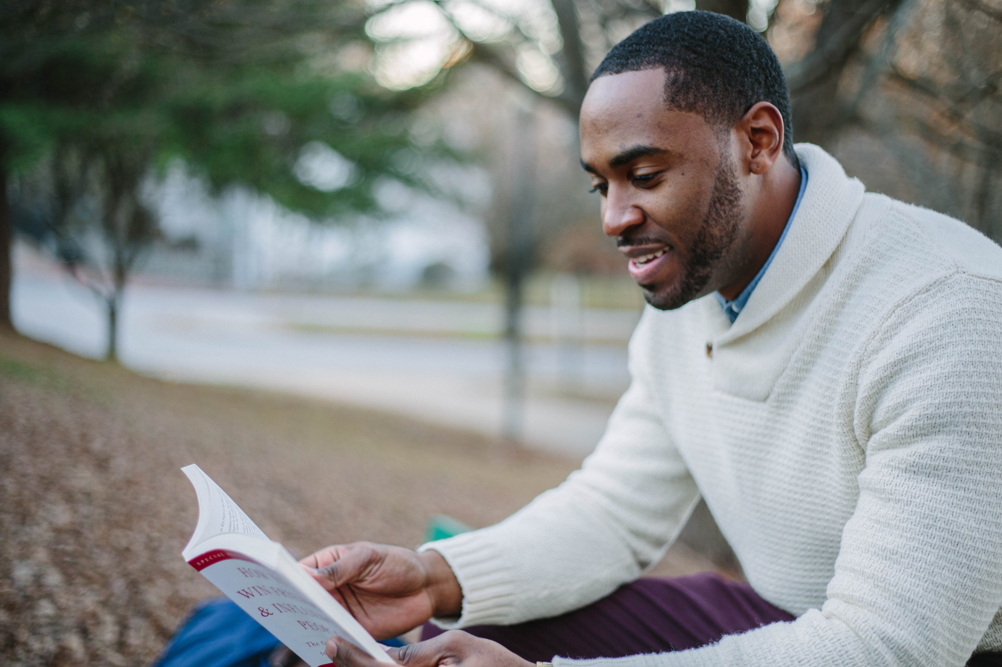 Black man wearing a white sweater sitting outside reading a book