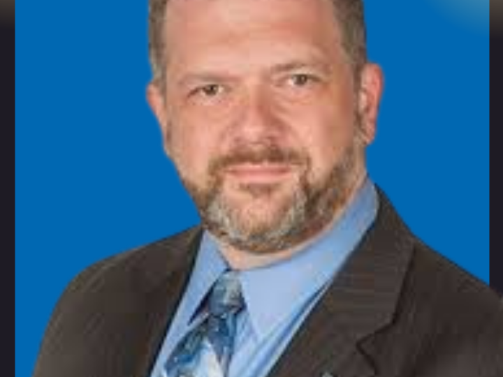 Headshot of a man with short brown hair and a beard, wearing a dark pinstriped suit, light blue shirt, and patterned blue tie. He is posed against a solid blue background.